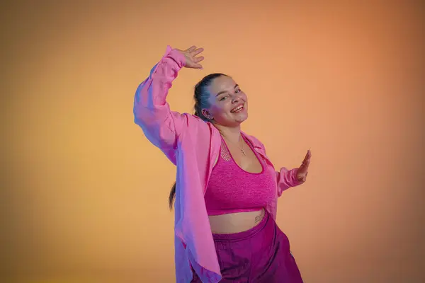 Young woman in a pink shirt dances to jazz funk rhythms against the backdrop of a orange studio. The girl confidently expresses her personality through dance, genuinely enjoying the music