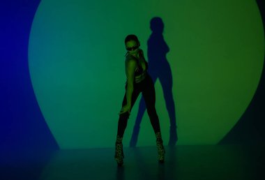 Genç bir kadın, spot ışıkları altında karanlık bir stüdyoda poz veriyor. Vücudunun gölgesi duvara yansıyor. Bir bayan dansçı yüksek topuklularla dans eder.