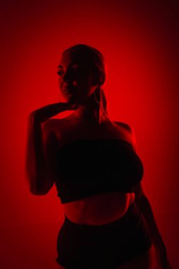 Siyah şortlu siluet kadın dansçı ve yüksek topuklularla dans ediyor. Genç kadın zarif bir şekilde poz veriyor ve karanlık stüdyoda kırmızı ışıkla ince, esnek bir vücut gösteriyor.