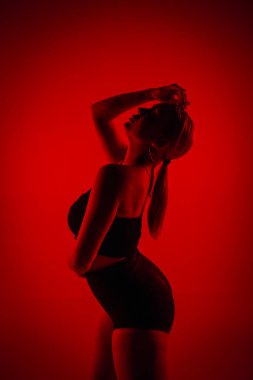 Siyah şortlu, siluetli kadın dansçı. Genç kadın zarif bir şekilde poz veriyor ve karanlık stüdyoda kırmızı ışıkla ince, esnek bir vücut gösteriyor. Yüksek topuklu elementler dans koreografisi