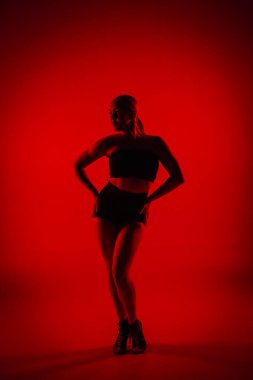 Siyah şortlu, siluetli kadın dansçı. Genç kadın zarif bir şekilde poz veriyor ve karanlık stüdyoda kırmızı ışıkla ince, esnek bir vücut gösteriyor. Yüksek topuklu elementler dans koreografisi