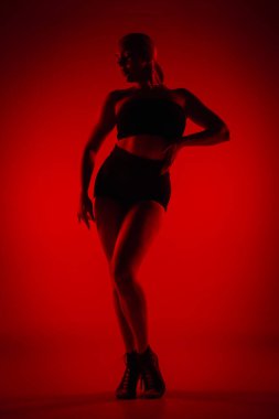 Siyah şortlu, siluetli kadın dansçı. Genç kadın zarif bir şekilde poz veriyor ve karanlık stüdyoda kırmızı ışıkla ince, esnek bir vücut gösteriyor. Yüksek topuklu elementler dans koreografisi