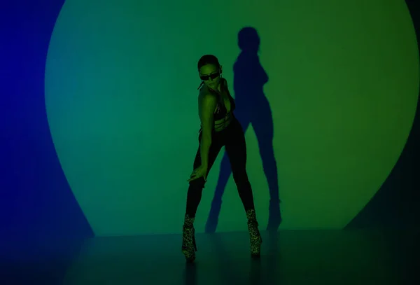 年轻女子在黑暗的演播室里摆姿势 蓝色的绿光作为聚光灯的中心 她身体的阴影反映在墙上 一个女舞蹈演员穿着高跟鞋表演舞蹈元素 — 图库照片