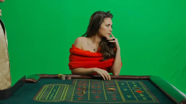 赌场和赌博商业广告的概念 漂亮的女性在工作室的彩色键绿色屏幕上 穿着红衣服的女人和蹲在轮盘赌桌旁的男人都在向一边望去 — 图库照片