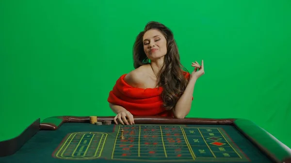 赌场和赌博商业广告的概念 漂亮的女性在工作室的彩色键绿色屏幕上 穿着红色衣服的女人坐在轮盘赌桌旁 开心地享受着这一表情 — 图库照片