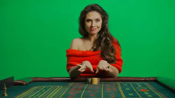 Casino and gambling commercial advertisement concept. Gorgeous female in studio on chroma key green screen. Appealing woman in red dress sitting at the roulette table smiling placing bet.