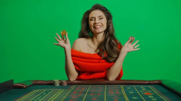 赌场和赌博商业广告的概念 漂亮的女性在工作室的彩色键绿色屏幕上 身穿红衣服的女人坐在轮盘赌桌旁笑着 手里拿着筹码 — 图库照片