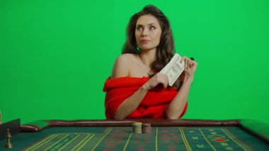 Kumarhane ve kumar yaratıcı reklam konsepti. Krom anahtar yeşil ekran yakından çekilmiş kadın portresi. Rulet masasında kırmızı elbiseli zarif bir kadın bir tomar para koyuyor. Yavaş çekim.