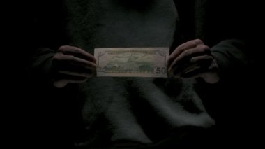 İş ve para yaratıcı reklam konsepti. Düşük ışıkta siyah arka planda izole edilmiş siyah elbiseli erkek portresi. Elinde elli dolarlık banknotla kameraya bakan adam..