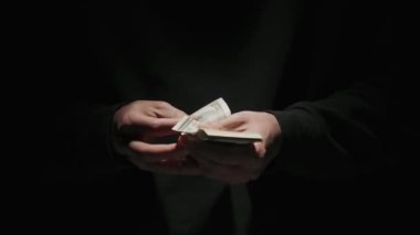 İş ve para yaratıcı reklam konsepti. Düşük ışıkta siyah arka planda izole edilmiş siyah elbiseli erkek portresi. Elinde bir tomar para tutan ve notlar sayan bir adam..