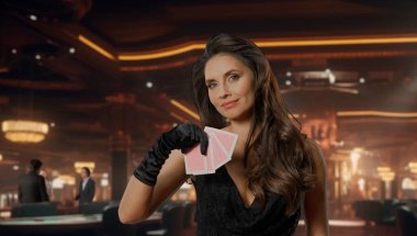 Kumarhanede 21 oynamak için poker masasında siyah elbiseli şık bir kadın. Poker kartları tutan, kameraya bakıp gülümseyen bir kadın. Kumarhane ve kumar kavramı