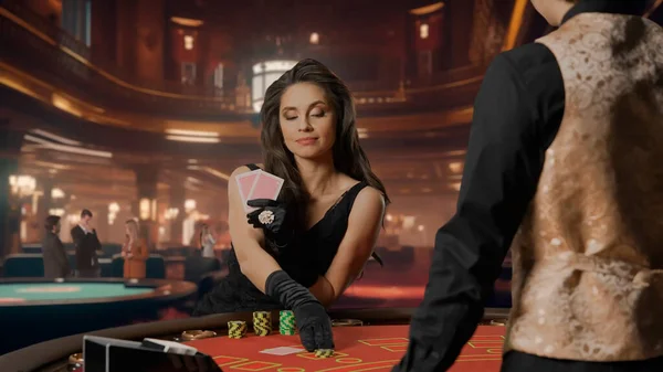 Elegant woman in black dress in casino. Attractive woman at blackjack poker table. Woman holding cards and betting with chips. Male croupier waiting for bet. Concept of casino and gambling
