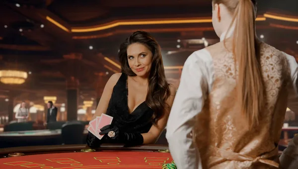 Attractive woman in black dress at poker table for blackjack game in casino. Woman with cards in hand smiles at the female croupier. Concept of casino and gambling