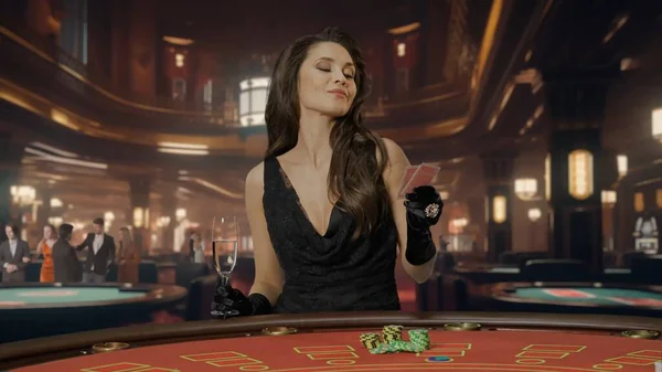 Chic woman in black dress at poker table for blackjack game in casino. The woman is holding two cards and a champagne glass. Concept of casino and gambling