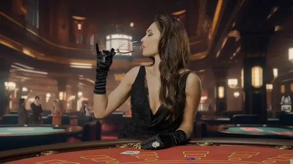 Chic woman in black dress at poker table for blackjack game in casino. The woman drinks champagne from a glass and celebrates her victory. The concept of casinos and gambling