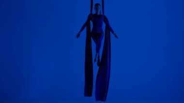 Modern koreografi ve akrobasi yaratıcı reklam konsepti. Mavi neon stüdyo arka planında izole edilmiş kadın sanatçı silueti. Kız dansçı hava ipeği üzerinde süzülüyor..