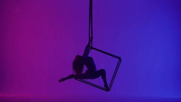 Modern choreography and acrobatics creative advertisement concept. Silhouette of female acrobat isolated on neon background. Woman aerialist dancer spinning in splits in the air on cube with ropes.