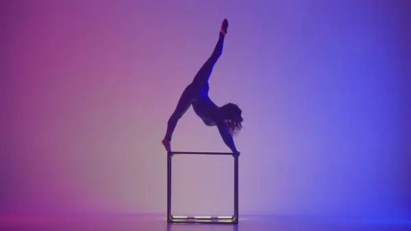 Modern choreography and acrobatics creative advertisement concept. Silhouette of female acrobat isolated on blue purple neon background. Woman gymnastic dancer in bodysuit performs on a cube.