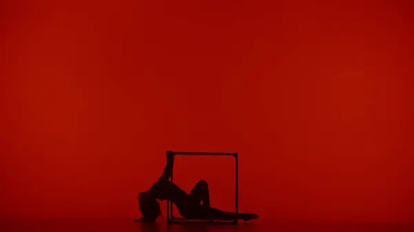 Modern choreography and acrobatics creative advertisement concept. Silhouette of female acrobat isolated on red neon background. Woman gymnastic dancer in bodysuit showing acrobatic element on a cube.