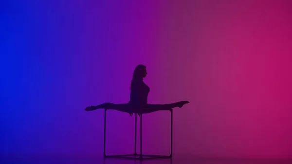 Modern choreography and acrobatics creative advertisement concept. Silhouette of female acrobat isolated on neon background. Woman gymnastic dancer in blue sparkling bodysuit showing split on a cube.