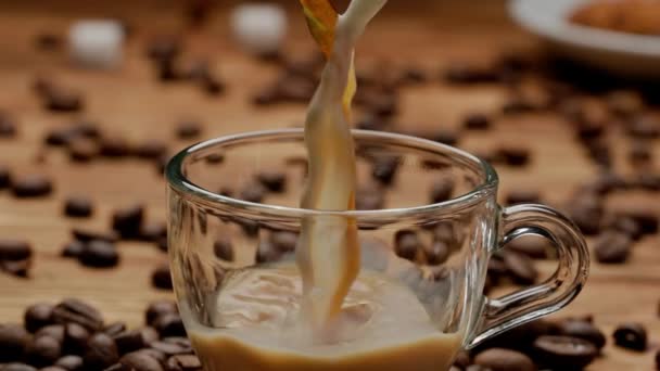 コーヒーとミルクのストリームがグラスカップに注ぎ込まれ 芳香族の飲み物で満たされます コーヒー豆の近くのキッチンテーブルのコーヒーまたはミルクティーのカップを閉じます 朝の儀式 朝食コンセプト — ストック動画