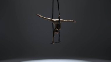 Modern koreografi ve akrobasi yaratıcı reklam konsepti. Kadın jimnastikçi monokrom stüdyosunda izole edilmiş. Akrobatik trapezdeki kız dansçı. Kayışları ikiye bölünmüş..