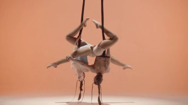 Modern koreografi ve akrobasi yaratıcı reklam konsepti. Kadın jimnastik düeti şeftali stüdyosunun arka planında izole edilmiş. Kız dansçılar başlarını jimnastik trapezine bağlı olarak eğiyorlar..