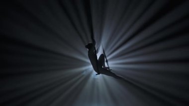Modern koreografi ve akrobasi yaratıcı reklam konsepti. Kadın jimnastikçi siyah arka plan ışınları ve dumanla izole edilmiş. Kız dansçı kız trapezde otururken kayışlarla element gösteriyor..