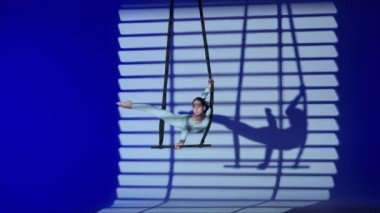 Modern koreografi ve akrobasi yaratıcı reklam konsepti. Kadın jimnastikçi, mavi neon stüdyo arka planında izole edilmiş. Kız dansçı kız kayışlarla akrobatik trapezde tutuyor ve dönüyor..
