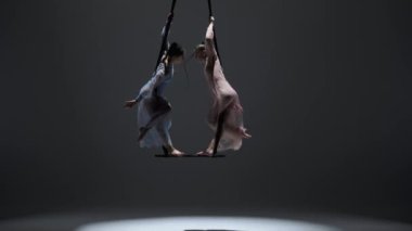 Modern koreografi ve akrobasi yaratıcı reklam konsepti. Kadın jimnastik ikilisi monokrom stüdyosunda izole edilmiş. Kızlar hava dansçıları kayışlarla akrobatik trapezde dönüyorlar..