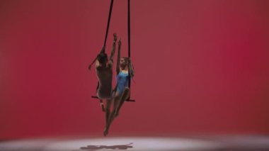 Modern koreografi ve akrobasi yaratıcı reklam konsepti. Kırmızı stüdyo arka planında izole edilmiş kadın jimnastik ikilisi. Kızlar hava dansçıları kayışlarla akrobatik trapezde oturup dönüyorlar..