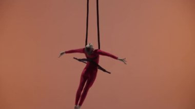 Modern koreografi ve akrobasi yaratıcı reklam konsepti. Turuncu stüdyo arka planında izole edilmiş kadın jimnastikçi. Kız hava dansçısı jimnastik kayışlarıyla trapezde asılı, hızla dönüyor..