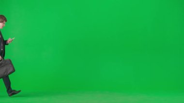 Modern iş adamı yaratıcı reklam konsepti. Krom anahtar yeşil ekranda erkek modelin portresi. Modayı takip eden takım elbiseli genç işadamı akıllı telefonu kontrol ediyor ve daha hızlı koşuyor. Yavaş çekim.