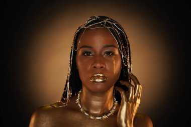 Saçında altın takılar olan Afro-Amerikan bir kadının portresi. Modellerin gözleri, dudakları ve elleri sıvı altınla kaplıdır. Kızların yanaklarından altın bir gözyaşı akıyor. Kapat..