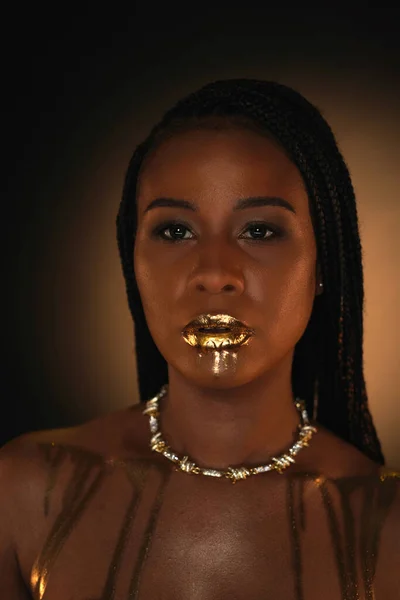 Portrait of an African American woman with a gold necklace around her neck. There are streaks of liquid gold on the models lips and collarbones. Close up. Beauty concept