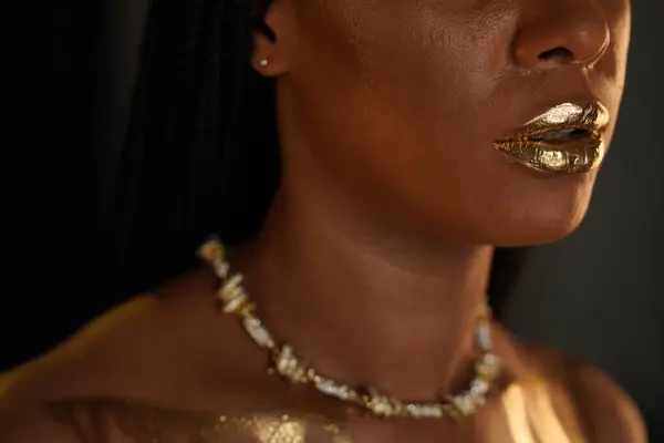 Portrait of an African American woman with a gold necklace around her neck. There are streaks of liquid gold on the models lips and collarbones. Close up. Beauty concept
