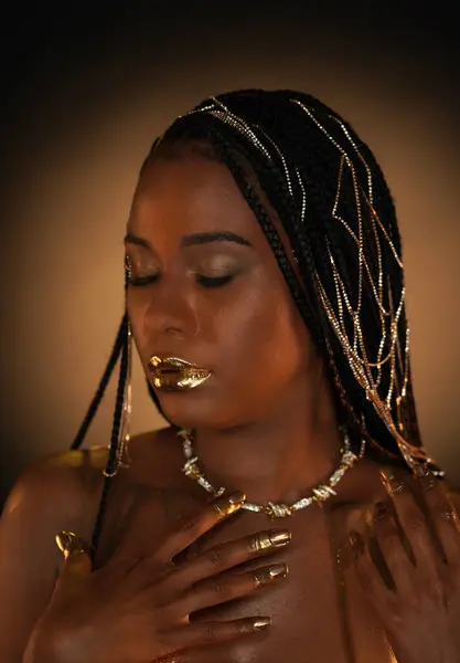 Portrait of an African-American woman with a gold necklace and gold chains in her hair. The models lips, fingers and nails are covered with liquid gold. Close up. The concept of beauty