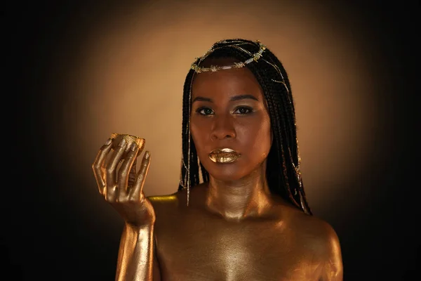Portrait of an African-American woman with gold jewelry in her hair. The models arms, neck and shoulders are covered with liquid gold. The girl is holding a golden lemon in her hands. Close up.