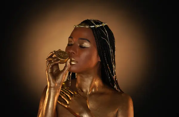 Portrait of an African-American woman with gold jewelry in her hair. The models arms, neck and shoulders are covered with liquid gold. The girl is holding a golden lemon in her hands. Close up.