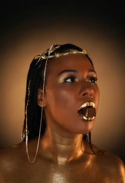 Portrait of an African-American woman with gold jewelry in her hair. The models arms, neck and shoulders are covered with liquid gold. The girl is holding a chocolate candy with her lips. Close-up.