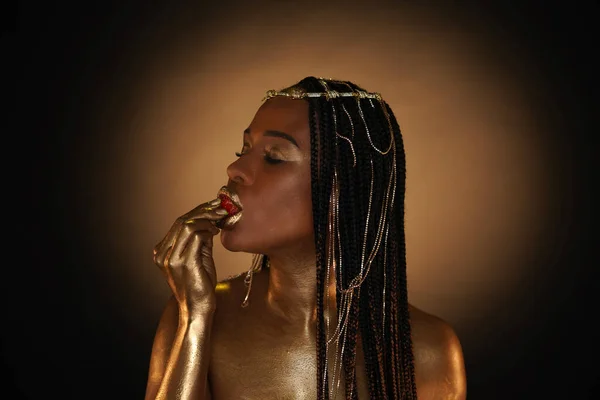 Portrait of an African-American woman with gold jewelry in her hair. The models arms, neck, lips and shoulders are covered with liquid gold. A girl is biting a strawberry. Closeup