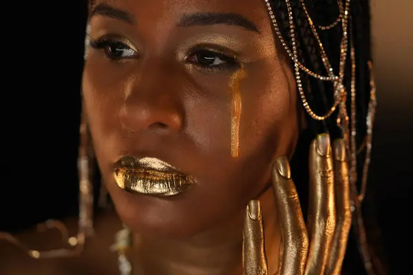 Portrait of an African-American woman with gold jewelry in her hair. The models eyes, lips and hands are covered with liquid gold. A golden tear is flowing down the girls cheek. Close up.