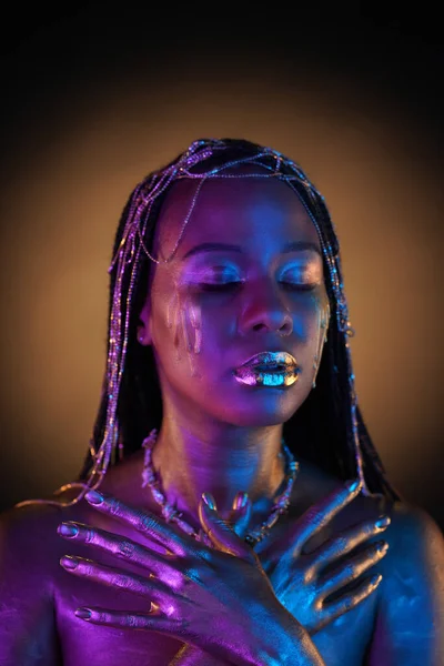 African American woman with gold jewelry in her hair. The models body covered with liquid gold glistens in blue violet neon light. Golden tears are flowing down the girls cheeks. Close up