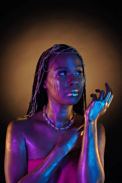 African American woman with gold jewelry in her hair. The models body covered with liquid gold glistens in blue violet neon light. Golden tears are flowing down the girls cheeks. Close up