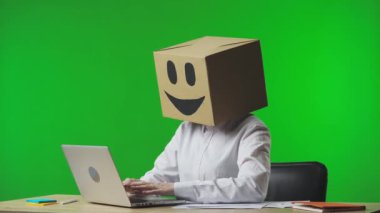 Stüdyonun yeşil arka planında gülümseyen emojisi olan mukavva kutudaki kadın. Çalışan mutlu bir şekilde dizüstü bilgisayarda klavye kullanıyor ve başarıyla seviniyor. İş hayatı ve günlük rutin işler.