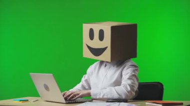 Stüdyonun yeşil arka planında gülümseyen emojisi olan mukavva kutudaki kadın. İşçi klavyeye yazı yazıyor ve sonra laptopunu kapatıyor. İş hayatı ve günlük rutin işler.