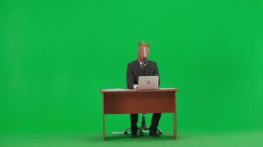 Atölye yeşil arka planda at başlı takım elbiseli bir adam. İş adamı masada oturuyor ve dizüstü bilgisayarda klavye çalıyor. Ağır ofis işi konsepti