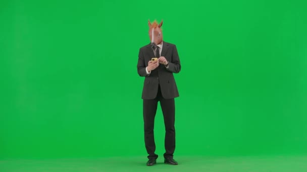 一个穿着西装 戴着马头面具 背景为绿色工作室的男人 一位商人在智能手机上看到某样东西 为好消息或胜利而感到高兴 艰苦工作概念 — 图库视频影像