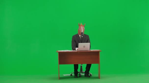 スタジオグリーンの背景に馬のヘッドマスクとビジネスススーツの男 ビジネスマンが机の上に座り ラップトップのキーボードに入力します ヘビーオフィスワークコンセプト — ストック動画