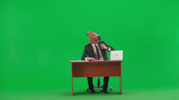 身穿西装 头戴马头面具 背景为工作室绿色 商人坐在办公桌前 说了些什么 然后把手指向两边 繁重工作的概念 — 图库视频影像